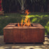 Lareira quadrada em aço corten com chamas acesas em jardim externo.