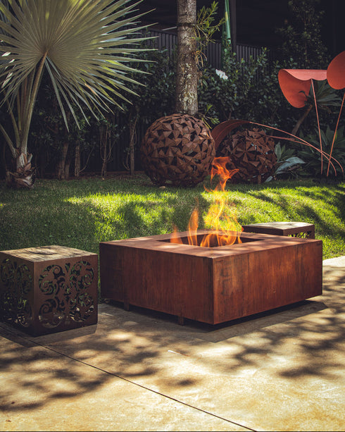 Lareira quadrada em aço corten acesa em jardim com decoração de esferas metálicas.