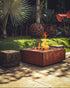 Lareira quadrada em aço corten acesa em jardim com decoração de esferas metálicas.